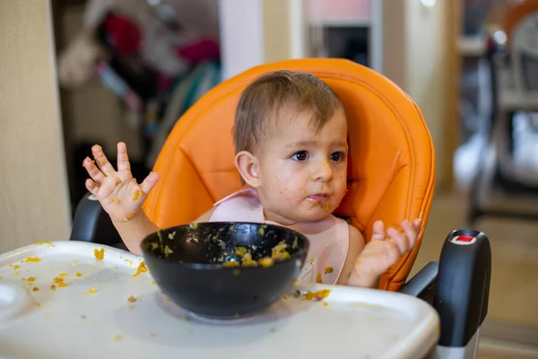Linda niña en un asiento de niño naranja delante de un plato de comida arroja sus manos y mira a los padres detrás de la cámara. todo alrededor en la comida — Foto de Stock