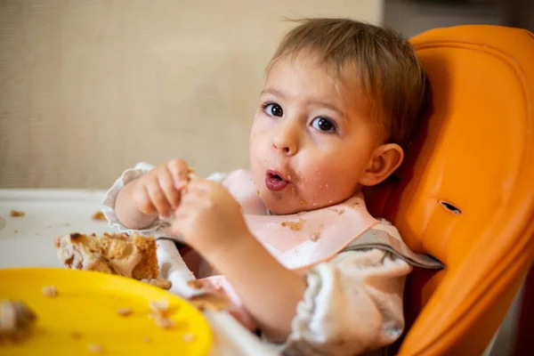Un bebé lindo está sentado en un asiento de niño naranja con una mesa con las manos y la cara sucias, mirando a la cámara y haciendo muecas. hay migajas y un plato naranja en la mesa. primer plano, vista frontal, enfoque suave . — Foto de Stock