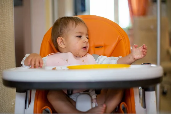Linda niña caucásica sentarse en silla de bebé naranja después de comer con migajas en la cara y mira a la mano en migajas de mesa y un plato de plástico naranja. primer plano, vista frontal, enfoque suave — Foto de Stock