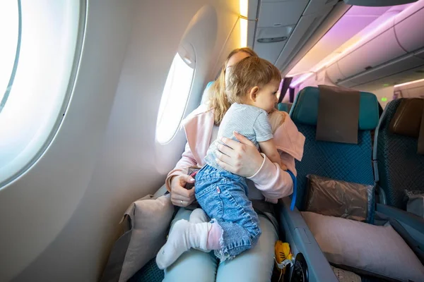 Concept of flight with a child. little cute toddler jumping on her knees with a young beautiful mother in an airplane chair — Stock Photo, Image