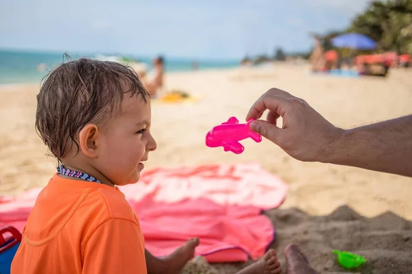 Koncepcja kwarantanny końcowej, zniesione ograniczenia, otwarte granice. Mały uroczy maluch siedzi na plaży, patrzy na zabawkowy samolot trzymany przez rodzica i śmieje się. — Zdjęcie stockowe