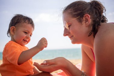 Küçük tatlı çocuk kumsalda annesiyle kum oynuyor. Arka planda güneşli bir günde ılık deniz