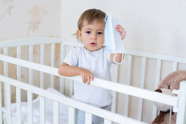 콘셉트 격리, 보안 위생. 작고 귀여운 아기는 집 요람에 있고 의료용 마스크를 들고 있습니다 — 스톡 사진