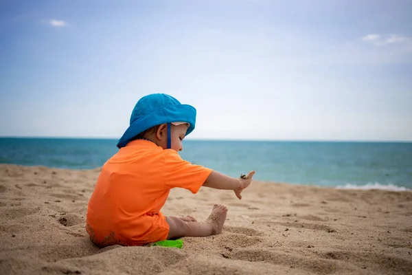 Koniec kwarantanny, otwarcie granic, swoboda podróżowania. maluch w niebieskim kapeluszu bawiący się piaskiem na plaży nad morzem. wakacje z dziećmi w pobliżu oceanu — Zdjęcie stockowe