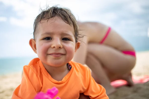 Portret małego słodkiego mokrego po kąpieli szczęśliwego i zmęczonego dzieciaka patrzącego w kamerę na tle piaszczystej plaży, zarazy i mamy — Zdjęcie stockowe