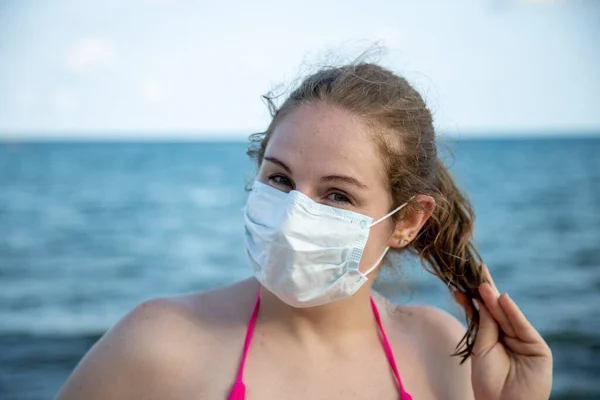 Retrato de una joven caucásica en una máscara médica y en un traje de baño mirando a la cámara alegremente. En el fondo el mar está borroso . — Foto de Stock