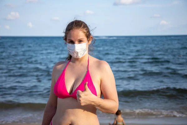 Joven chica caucásica en una máscara médica y en un traje de baño mira a la cámara y muestra el pulgar hacia arriba. En el fondo, en la playa junto al mar, un niño juega borroso — Foto de Stock