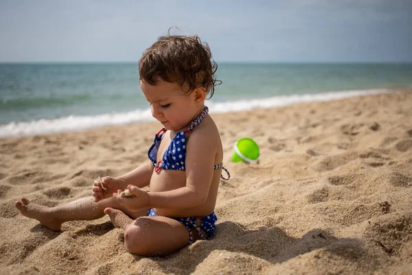 Mała słodka dziewczynka maluch w niebieskim stroju kąpielowym na piaszczystej plaży studiuje piasek na tle fal morskich w ciepły słoneczny dzień — Zdjęcie stockowe