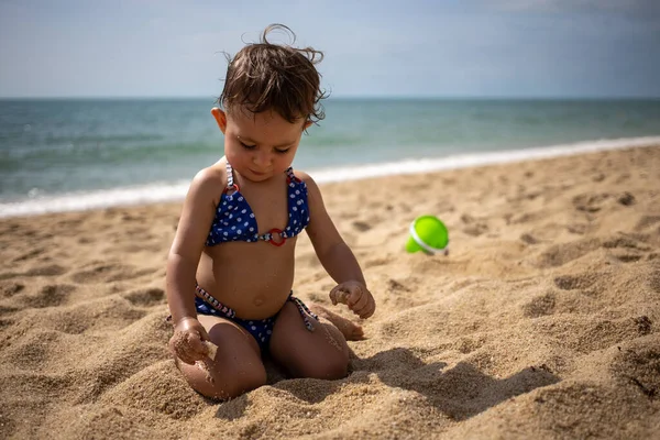 Mała słodka dziewczynka maluch w niebieskim stroju kąpielowym na piaszczystej plaży bawi się w piasek na tle fal morskich w ciepły słoneczny dzień — Zdjęcie stockowe