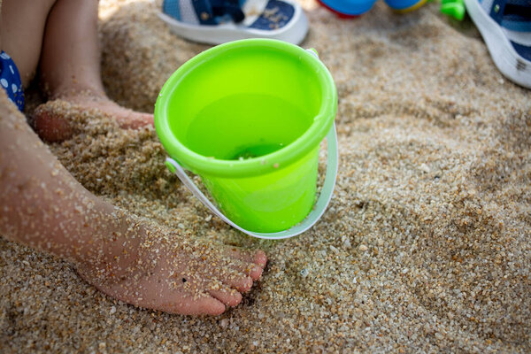 зеленый пластиковый ведро с водой стоит на песке на пляже у ног ребенка играет песок, крупным планом
