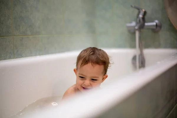 Крихітне кавказьке немовля приймає ванну, грайливе в ванній і посміхається, дивлячись на сплески води, на задньому плані зелена ванна в тумані. М'який фокус. — стокове фото