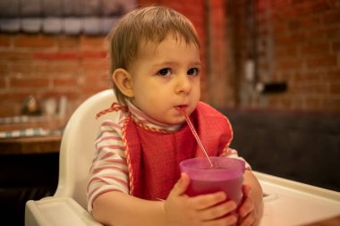 Sevimli bir çocuk sandalyeye oturur, elinde bir bardak meyve suyu tutar ve pipetten içer. Arka planda tuğladan duvarları olan rahat bir oda.