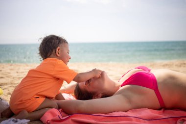 Küçük tatlı çocuk kumsalda yatan beyaz bir anneyle oynuyor ve onu rahatsız ediyor. Arka planda güneşli bir günde ılık deniz.