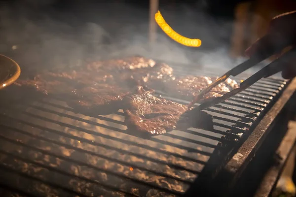 La sera un barbecue su cui vengono grigliate gustose bistecche succose su un fuoco aperto, una mano tiene le pinze e gira la carne. primo piano, messa a fuoco morbida. il fumo è evidenziato — Foto Stock