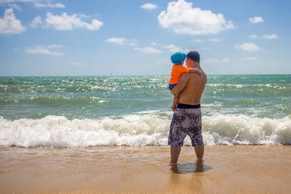 幼児の概念で旅をします。父は腕に幼児を抱えて砂浜に立つ波を見て — ストック写真