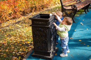 Akıllı nesil konsepti. Parktaki siyah konteynırın yanında duran iki küçük şirin bebek ve oraya çöp atıyorlar. Arkadan başka bir çocuk geliyor. yumuşak odak, bulanık arkaplan.