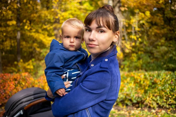 Retrato de uma jovem mãe bonita segurando uma criança em seus braços, vestida com um olhar de família azul sorrindo e olhando para a câmera n parque de outono — Fotografia de Stock