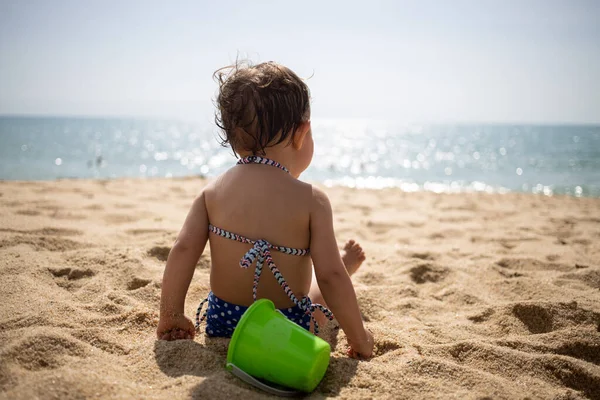 Filozofia dzieci. Little cute maluch opalony kaukaska dziewczyna w niebieskim stroju kąpielowym siedzi na piaszczystej plaży na tle fal morskich w ciepły słoneczny dzień. widok z tyłu, zbliżenie — Zdjęcie stockowe