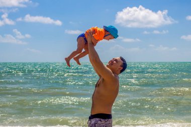 Babalar günü. Mutlu baba, neşeli küçük bir bebeği başının üstünde tutuyor. Kumlu deniz kıyısının arka planı