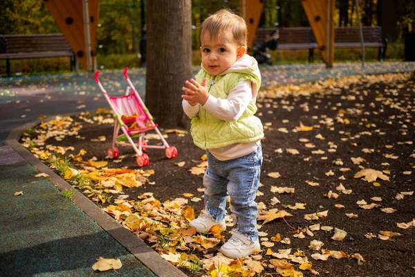 Linda niña de un año de edad, de pie en hojas amarillas y quitándose los bolígrafos y sonriendo alegremente a la cámara en el parque de otoño — Foto de Stock