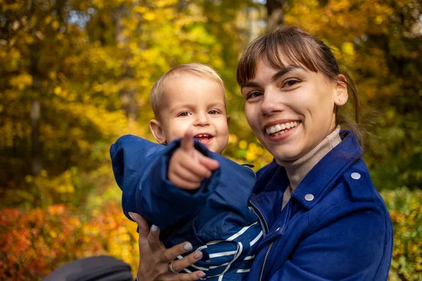 Jeune belle maman et tout-petit sur ses mains regarder la caméra et rire heureux dans le parc d'automne — Photo