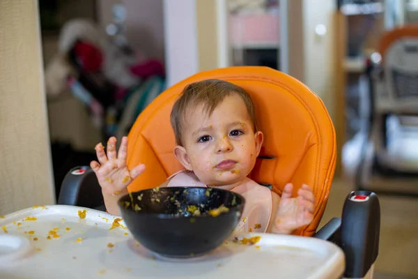 Linda niña en un asiento de niño naranja delante de un plato de comida arroja sus manos y mira a la cámara. por todas partes en migas de comida. primer plano, vista frontal, enfoque suave, fondo borroso — Foto de Stock
