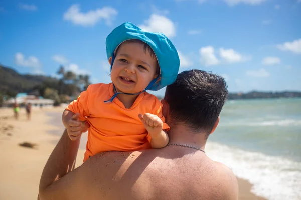 Pequeno feliz menino bonito sentado em seu pai ombro no fundo da praia e mar azul — Fotografia de Stock