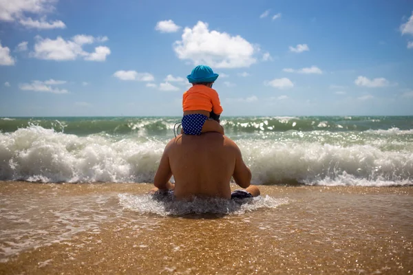 Tata z dzieckiem na ramionach. Spójrz na fale siedzące na plaży w falach. — Zdjęcie stockowe