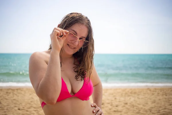 Chica rubia sonriendo y mirando juguetonamente a la cámara, vestida con un bikini rosa y corazones de gafas — Foto de Stock