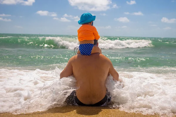 Dzień ojca. Obrońca taty. Dziecko siedzi na ramionach taty, tata siedzi na plaży i nadchodzi fala. — Zdjęcie stockowe