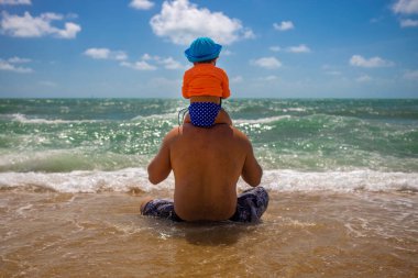 Babalar günü. Baba ve bebek sahilde oturur ve dalgaların arkasına bakar.