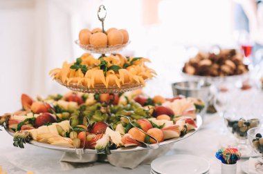 Meyve rafı: üzüm, elma, şeftali, ananas. Resepsiyon, yemek servisi, temiz bulaşıklar. Masa beyaz bir masa örtüsüyle kaplıdır..