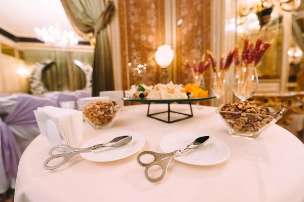 Kitchen tongs on plates. Shelves with assorted cheese, nuts, snacks on sticks in a glass. Reception, catering, clean dishes, napkins. The table is covered with a white tablecloth.
