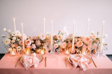 Ziyafet, restoran. Masa ayarı. Pembe tabaklar, altın çatal bıçak takımı, gözlük. Masanın üzerinde pembe bir masa örtüsü var. Altın yüzüklü şeftali renkli peçete. Şeftali pembesi renklerinde çiçek aranjmanları. Uzun mumlar.