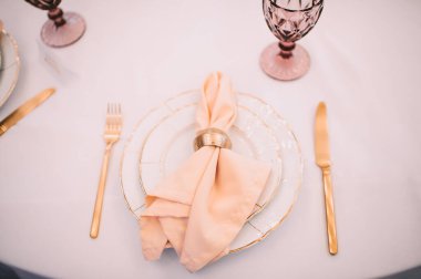 Ziyafet, restoran. Masa ayarı. Altın kenarlı beyaz tabaklar, altın çatal bıçak takımı, gözlük. Altın yüzüklü şeftali renkli peçete.. 