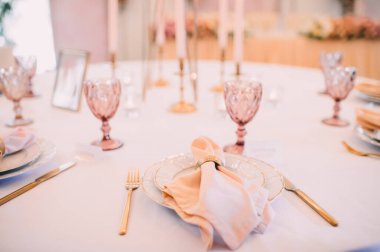 Ziyafet, restoran. Masa ayarı. Altın kenarlı beyaz tabaklar, altın çatal bıçak takımı, gözlük. Altın yüzüklü şeftali renkli peçete.. 