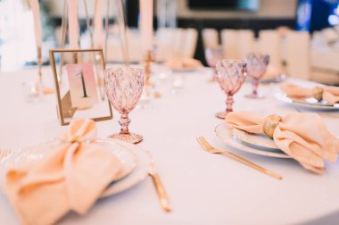 Ziyafet, restoran. Masa ayarı. Altın kenarlı beyaz tabaklar, altın çatal bıçak takımı, gözlük. Altın yüzüklü şeftali renkli peçete.. 