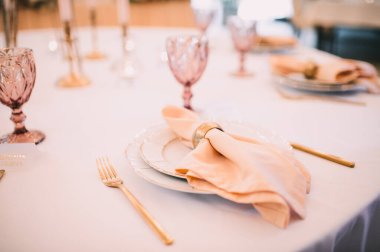 Ziyafet, restoran. Masa ayarı. Altın kenarlı beyaz tabaklar, altın çatal bıçak takımı, gözlük. Altın yüzüklü şeftali renkli peçete.. 