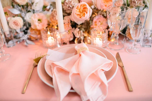 Banquet, restaurant. Table setting. Pink plates, gold cutlery, glasses. On the table is a pink tablecloth. Peach-colored napkin with a golden ring. Floral arrangements in peach pink hues. Tall candles