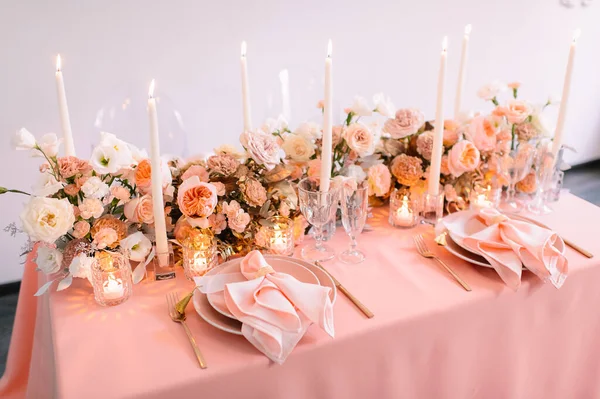 Banquet, restaurant. Table setting. Pink plates, gold cutlery, glasses. On the table is a pink tablecloth. Peach-colored napkin with a golden ring. Floral arrangements in peach pink hues. Tall candles