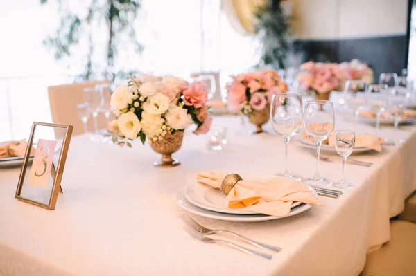 Banquet, restaurant. Table setting. White plates with a gold rim, golden cutlery, glasses. Peach-colored napkin with a golden ring. Peach pink floral arrangements