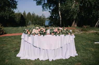 Yeşil çimenlikte ziyafet masası. Beyaz tahta sandalyeler. Çatal bıçak takımı. Çiçeklerin çiçek aranjmanı. Masanın üzerinde beyaz bir masa örtüsü var..