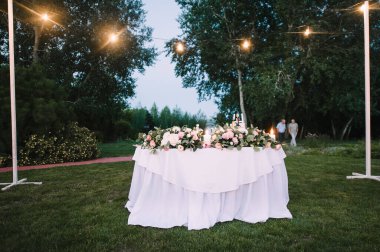 Yeşil çimenlikte ziyafet masası. Beyaz tahta sandalyeler. Çatal bıçak takımı. Çiçeklerin çiçek aranjmanı. Masanın üzerinde beyaz bir masa örtüsü var..