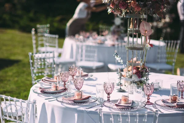 Banquet table on a green lawn. Racks and cutlery, velvet napkins, pink glasses. Floral arrangement of pink flowers. Silver candlesticks and candles. On the table is a white tablecloth.