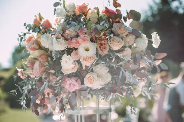 Banquet table on a green lawn. Racks and cutlery, velvet napkins, pink glasses. Floral arrangement of pink flowers. Silver candlesticks and candles. On the table is a white tablecloth.