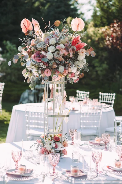 Banquet table on a green lawn, dance floor. Racks and cutlery, velvet napkins, pink glasses. Floral arrangement of pink flowers. Silver candlesticks and candles. On the table is a white tablecloth.