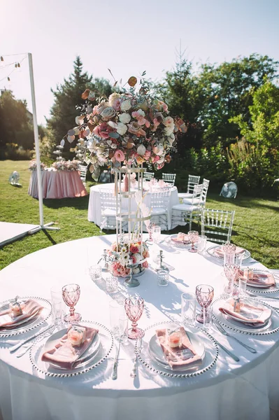 Banquet table on a green lawn, dance floor. Racks and cutlery, velvet napkins, pink glasses. Floral arrangement of pink flowers. Silver candlesticks and candles. On the table is a white tablecloth.