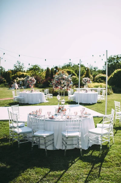 Banquet table on a green lawn, dance floor. Racks and cutlery, velvet napkins, pink glasses. Floral arrangement of pink flowers. Silver candlesticks and candles. On the table is a white tablecloth.