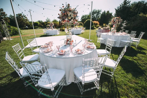 Banquet table on a green lawn, dance floor. Racks and cutlery, velvet napkins, pink glasses. Floral arrangement of pink flowers. Silver candlesticks and candles. On the table is a white tablecloth.