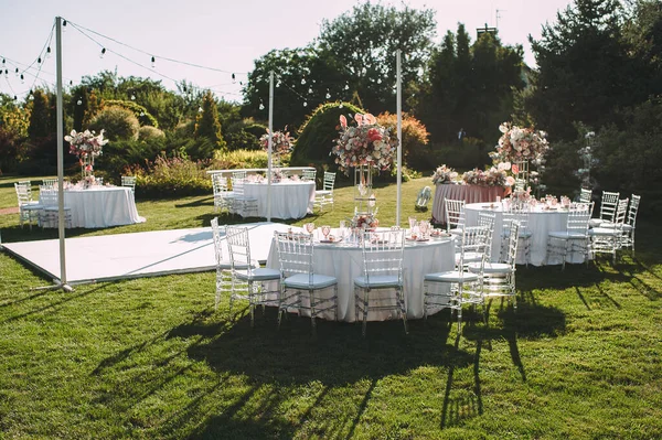 Banquet table on a green lawn, dance floor. Racks and cutlery, velvet napkins, pink glasses. Floral arrangement of pink flowers. Silver candlesticks and candles. On the table is a white tablecloth.